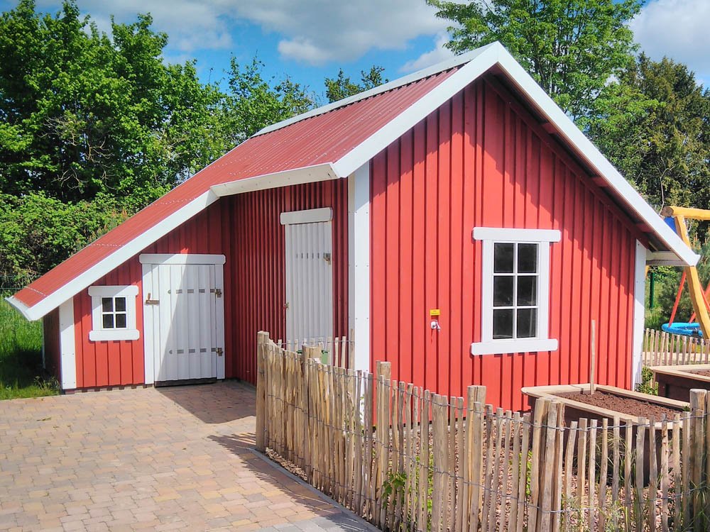 Spielhaus im skandinavischen Stil für Kindergarten und Spielplatz Rotes Holzspielhaus mit weißen Fenstern und Türen auf naturnahem Außengelände